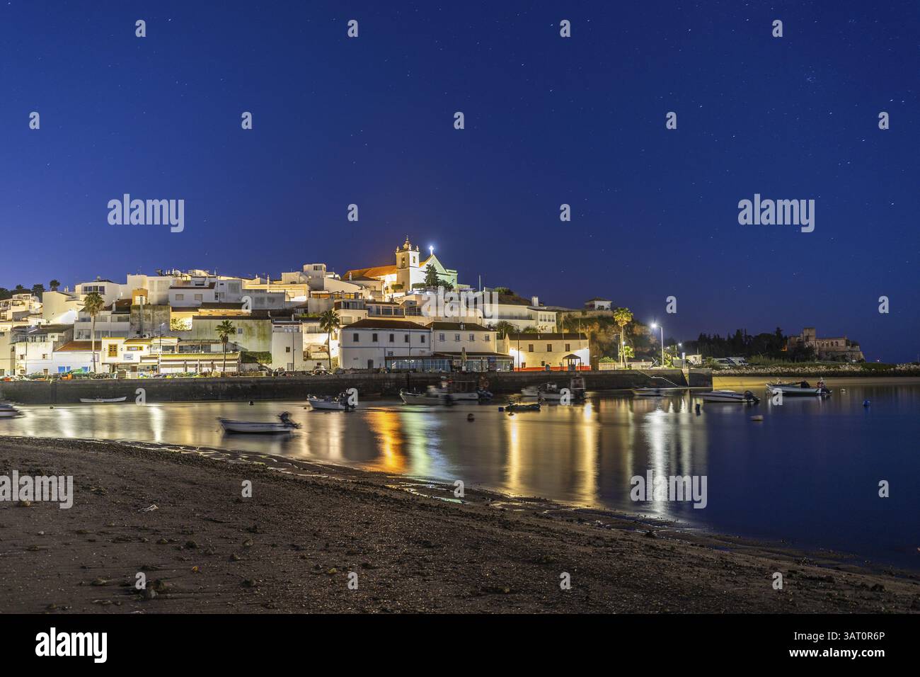 Nachtaufnahme von einem Sandstrand und Hafen bei Ebbe. Panoramabild eines beleuchteten historischen Stadtzentrums bei Sonnenaufgang in Ferragudo, Portimao, Algarve, Stockfoto