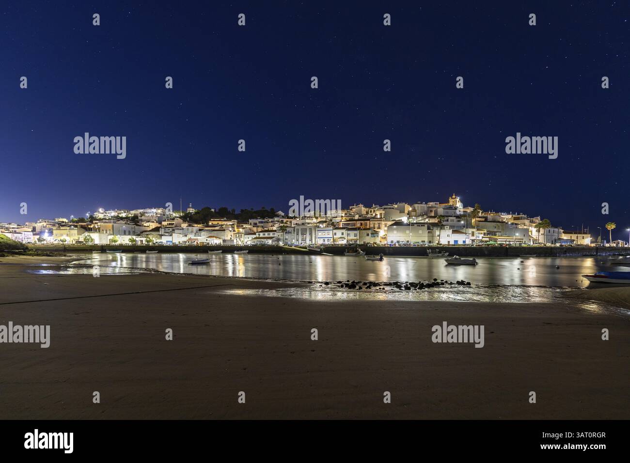 Nachtaufnahme von einem Sandstrand und Hafen bei Ebbe. Panoramabild eines beleuchteten historischen Stadtzentrums bei Sonnenaufgang in Ferragudo, Portimao, Algarve, Stockfoto