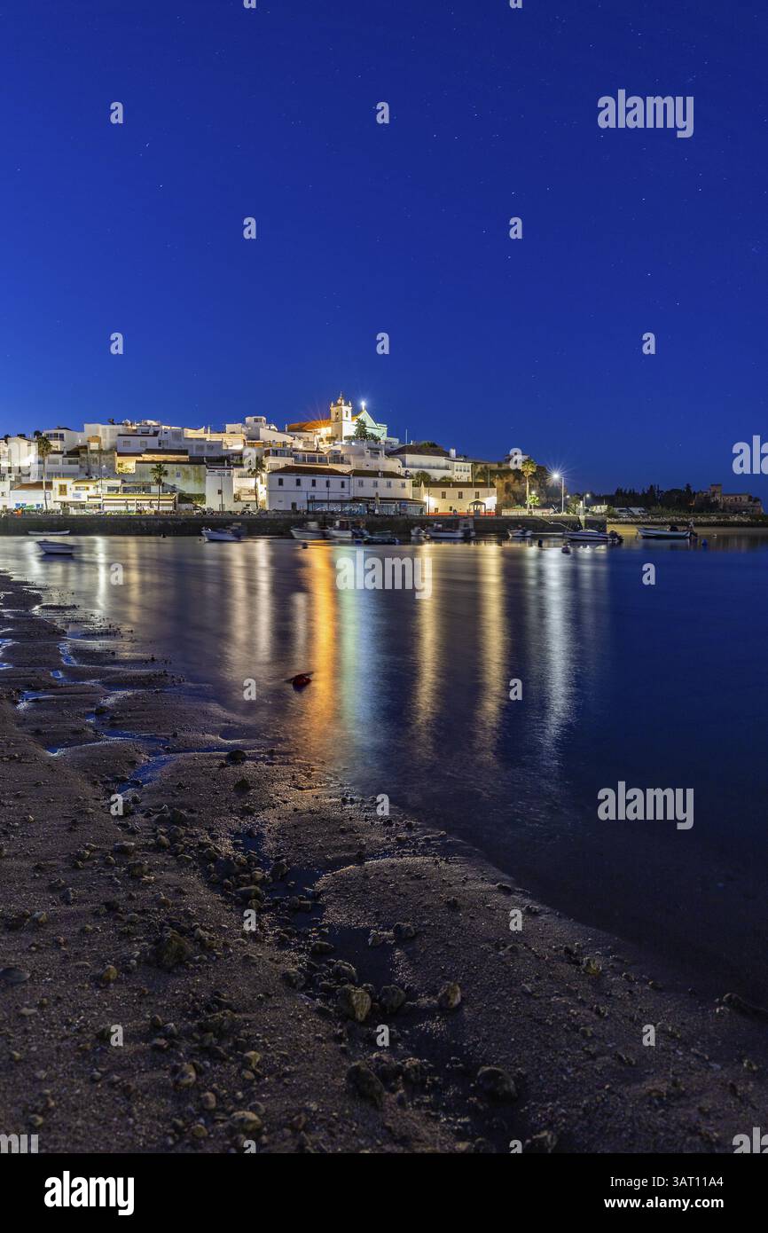 Nachtaufnahme von einem Sandstrand und Hafen bei Ebbe. Panoramabild eines beleuchteten historischen Stadtzentrums bei Sonnenaufgang in Ferragudo, Portimao, Algarve, Stockfoto