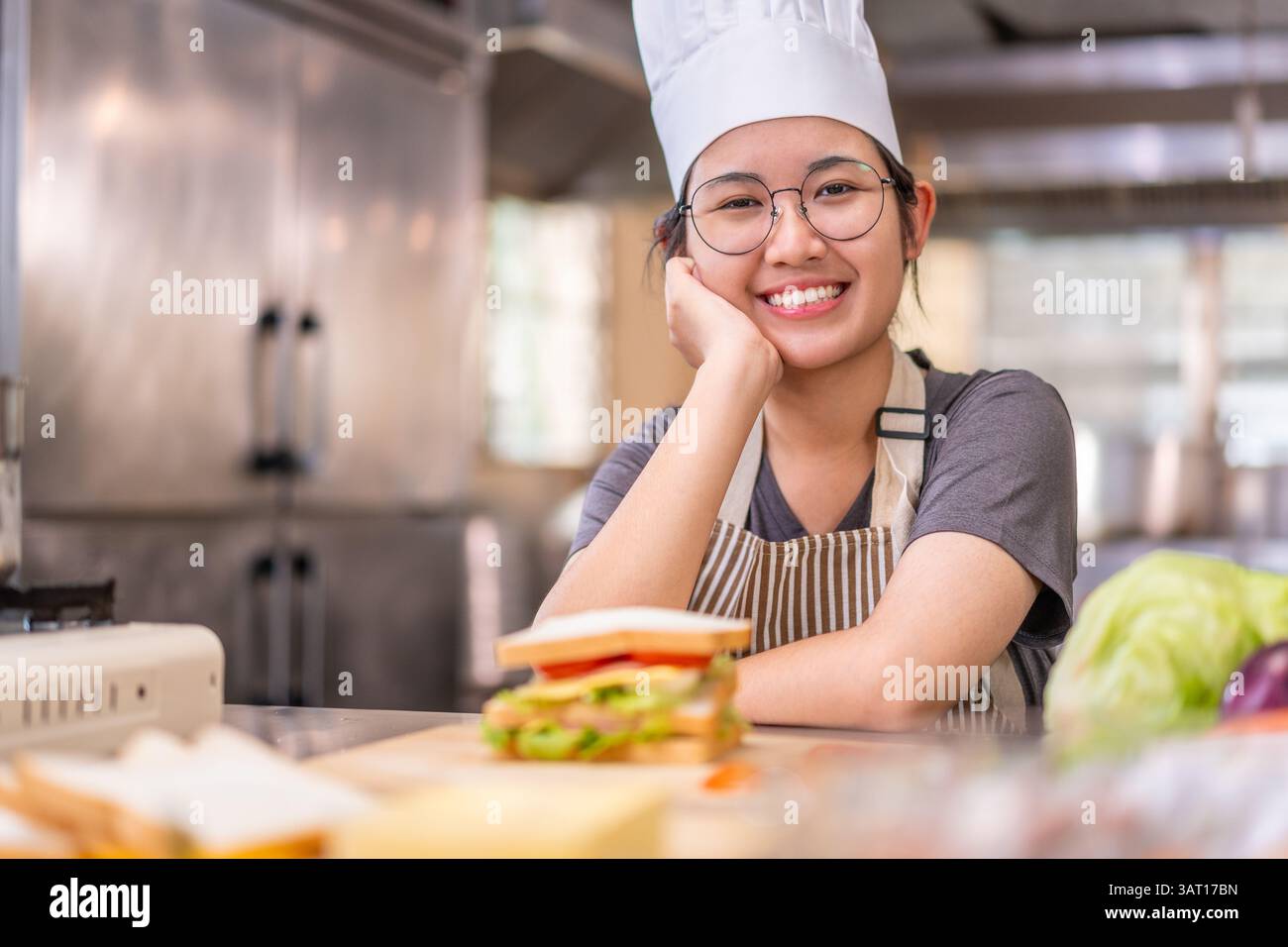 Eine fröhliche junge Küchenchefin mit weißem Hut und gestreifter Schürze lächelt, während sie ihren Kopf in einer kommerziellen Küche auf ihre Hand legt. Vor ihr Stockfoto
