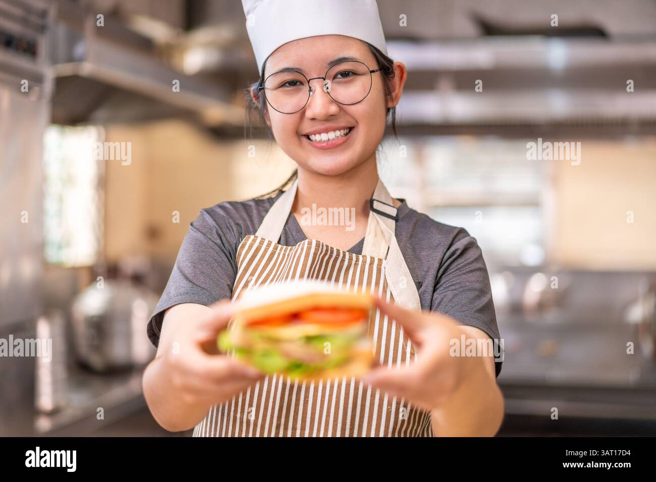 Eine fröhliche junge Küchenchefin mit gestreifter Schürze und weißem Kochhut präsentiert stolz ein frisch zubereitetes Sandwich in einem professionellen K vor die Kamera Stockfoto