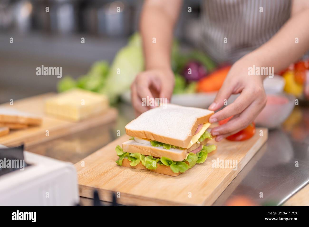 Nahaufnahme einer Person, die ein frisches Sandwich mit Salat, Käse, Ei und Fleischscheiben auf einem hölzernen Schneidebrett zusammenstellt. Umgeben von frischem Essen Stockfoto