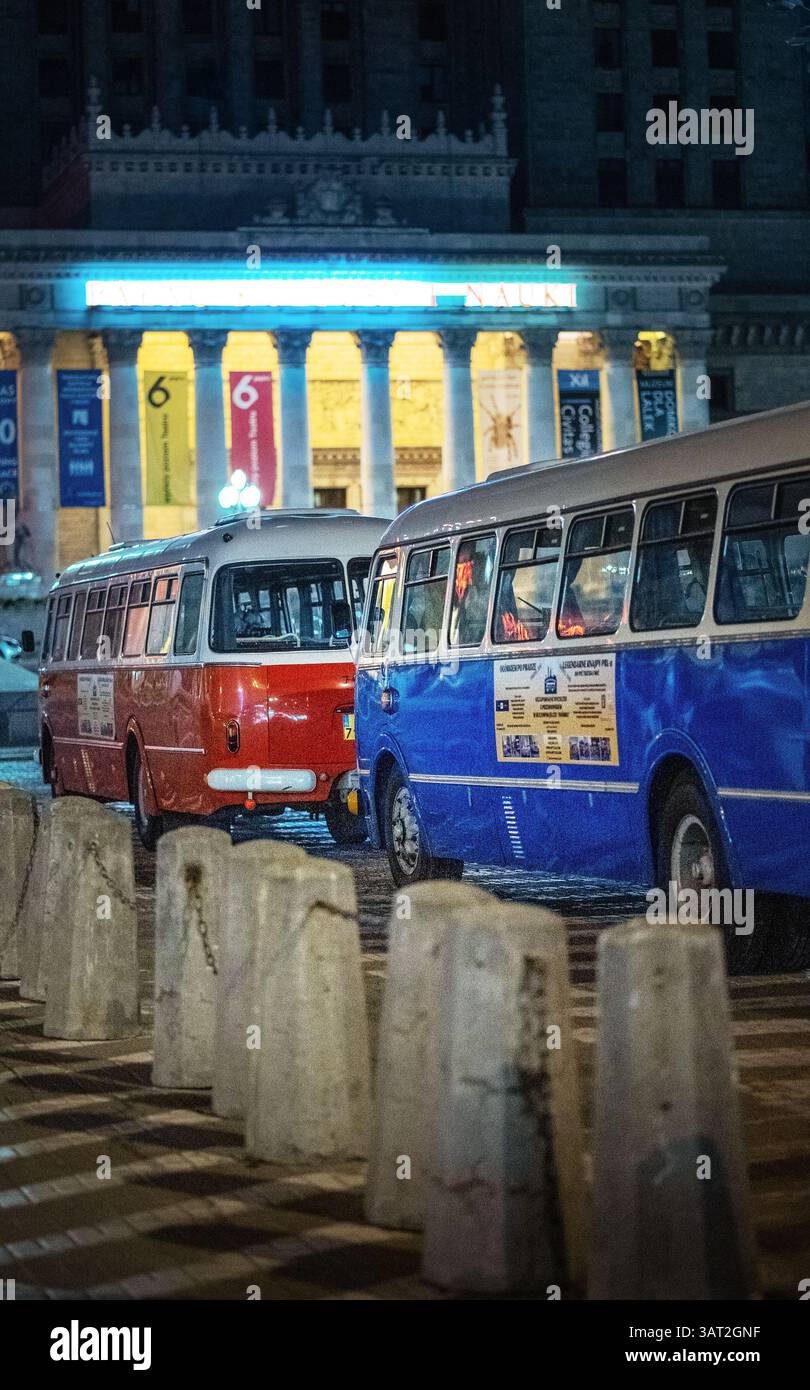 Zwei Oldtimer-Busse, einer orange und einer blau, parken nachts vor einem historischen Gebäude. Die Szene wird durch bunte Lichter beleuchtet, wodurch ein n entsteht Stockfoto