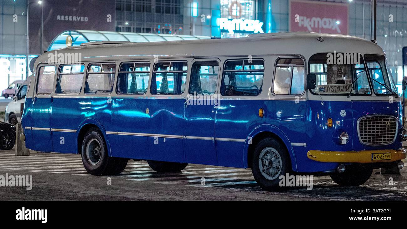 Ein klassischer blau-weißer Bus, der nachts auf einer Stadtstraße parkt und von Stadtlichtern beleuchtet wird. Der Bus hat ein klassisches Design mit großen Fenstern und einem Stockfoto