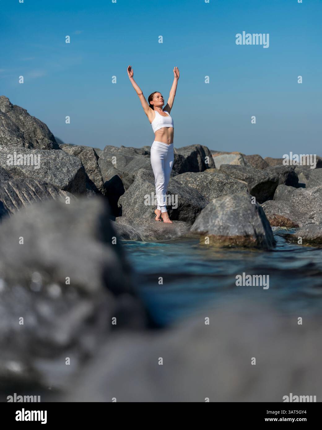 Sportliche Frau, die mit erhobenen Armen auf Felsen am Wasser steht Stockfoto