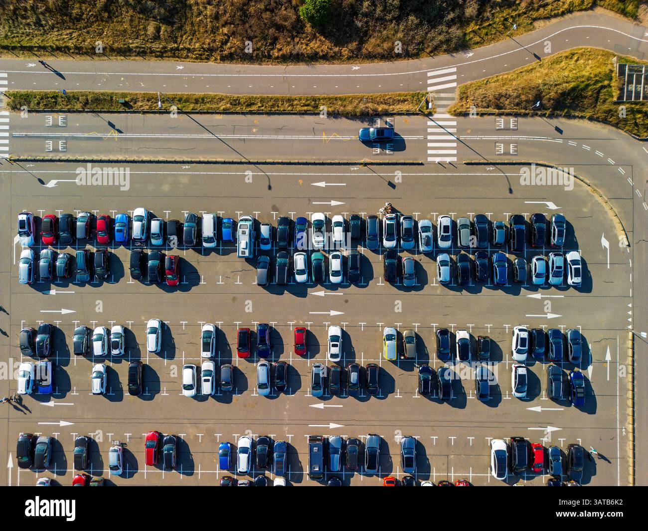 Aus der Vogelperspektive auf einen großen, organisierten Parkplatz voller farbenfroher Autos. Helles Sonnenlicht wirft lange Schatten und hebt die ordentlichen Reihen und Straßenmarkierungen hervor Stockfoto