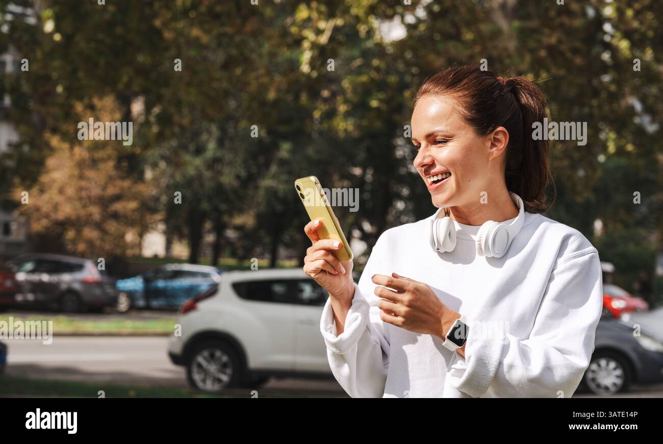 Junge Frau in weißem, lässigem Sweatshirt mit Smartphone auf der Stadtstraße und Kopfhörern um den Hals Stockfoto