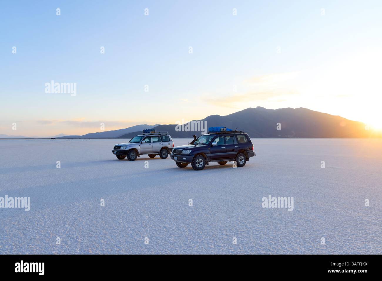 Zwei Geländewagen parkten bei Sonnenuntergang auf der strahlend weißen Salar de Uyuni Salzplatte. Toyota Land Cruiser mit Dachausrüstung vor Bergkulisse in B Stockfoto