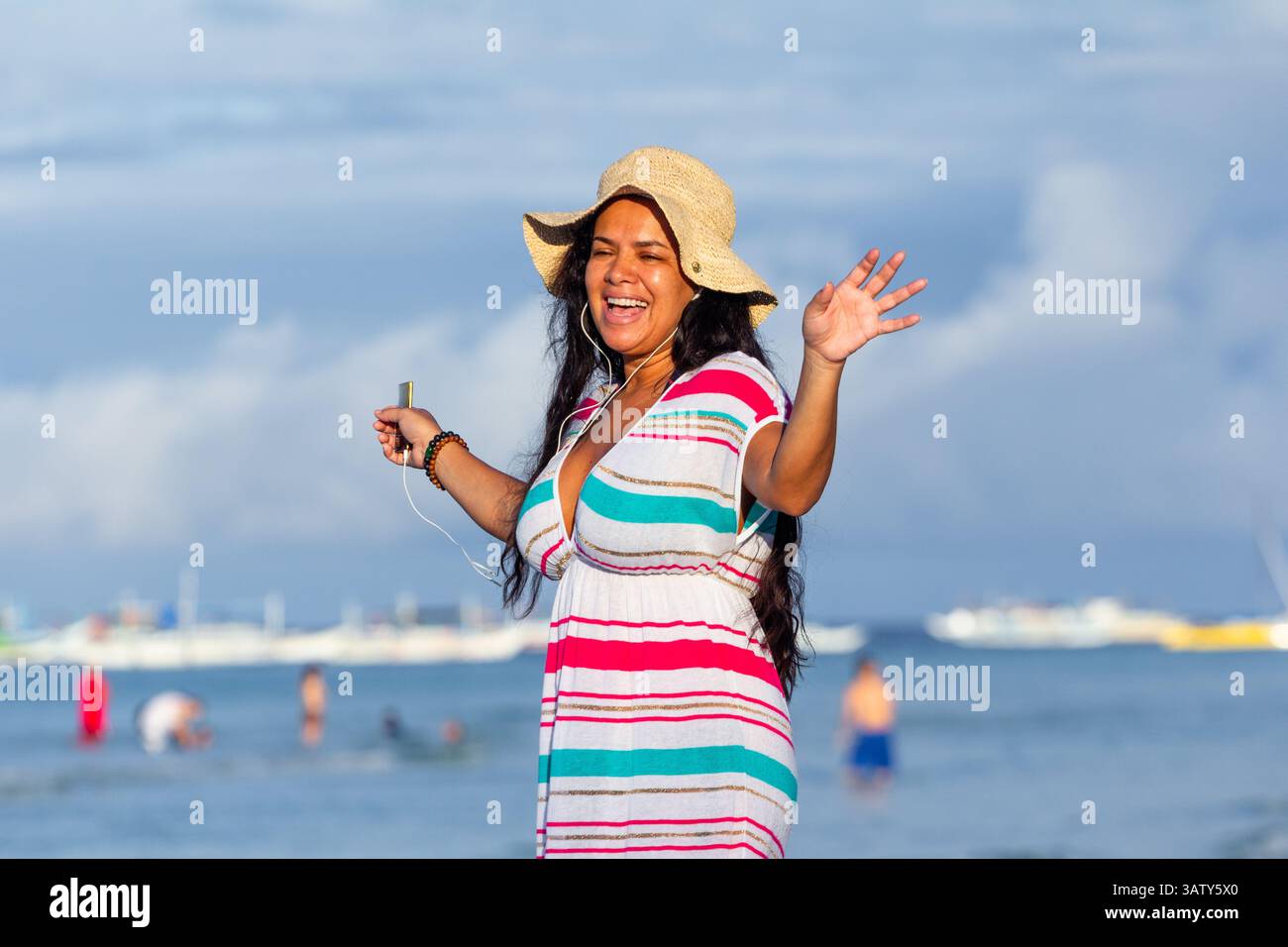 Eine Latina in Übergröße mit Krempenhut und buntem Kleid tanzt und singt fröhlich mit ihrem iPod Nano am malerischen Strand von Boracay Island Stockfoto