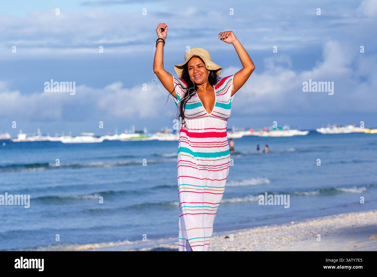 Eine Latina in Übergröße mit Krempenhut und buntem Kleid tanzt und singt fröhlich mit ihrem iPod Nano am malerischen Strand von Boracay Island Stockfoto