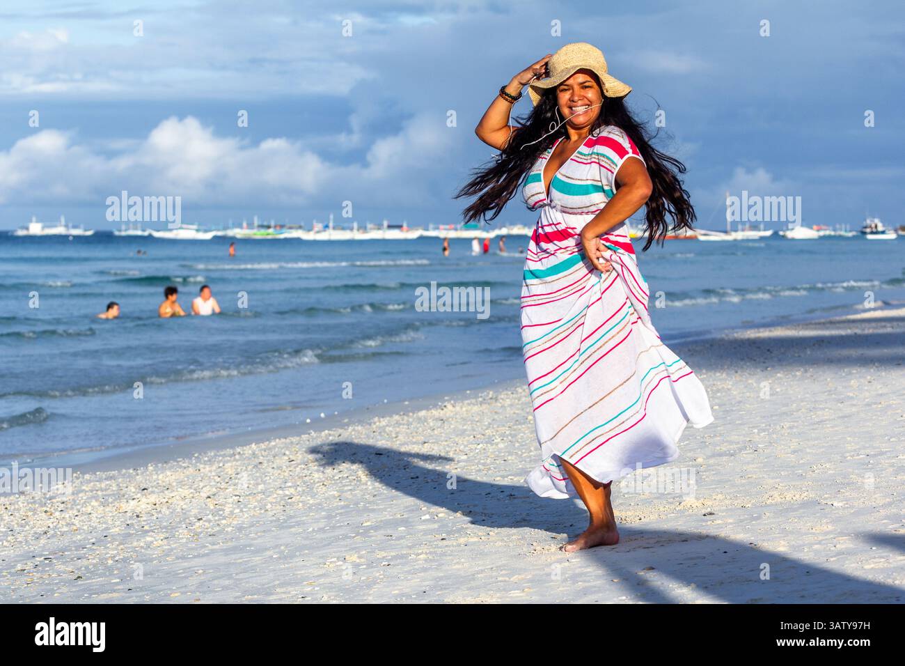 Eine Latina in Übergröße mit Krempenhut und buntem Kleid tanzt und singt fröhlich mit ihrem iPod Nano am malerischen Strand von Boracay Island Stockfoto