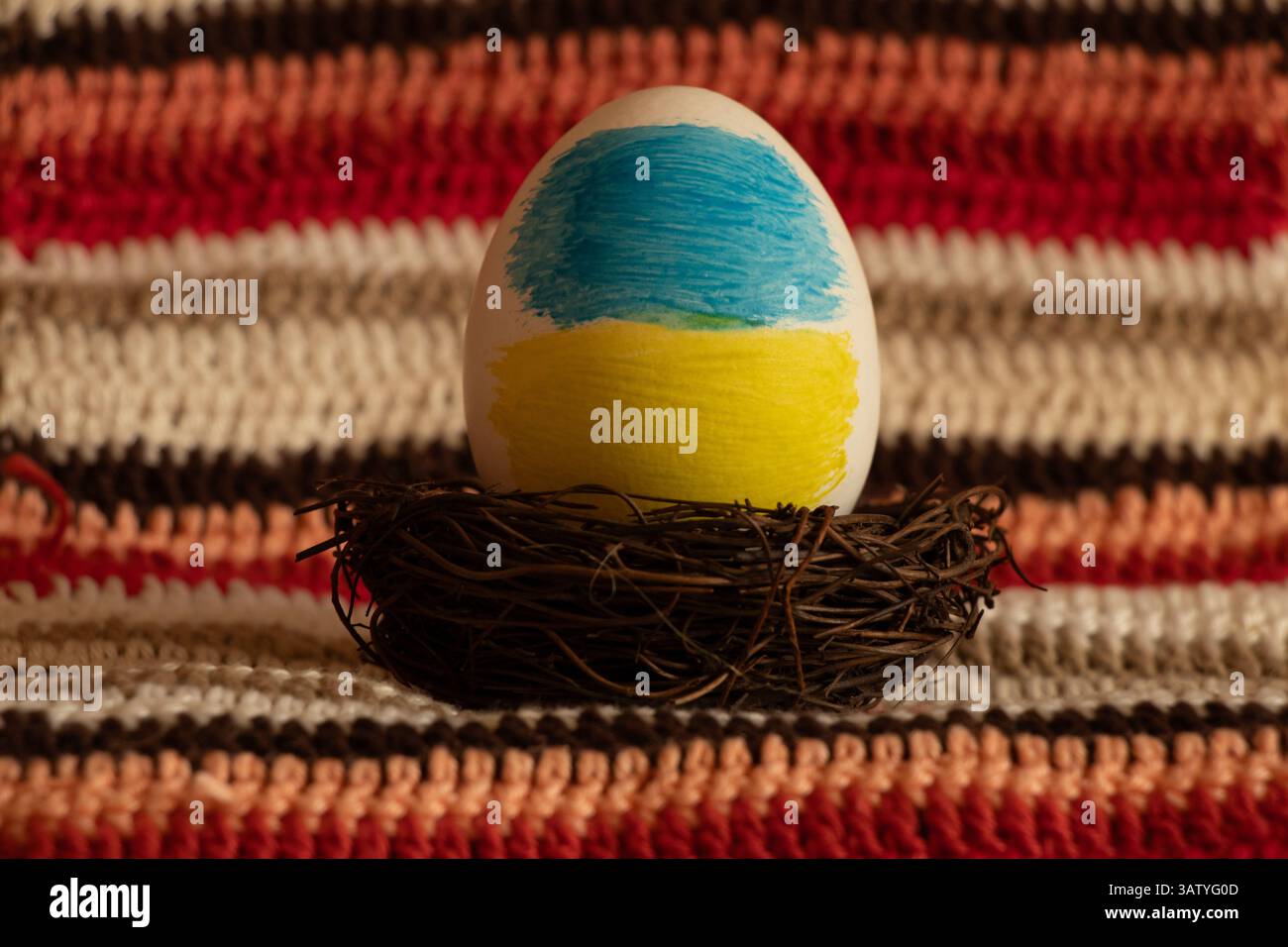 Ein Osterei in den Farben der ukrainischen Flagge befindet sich in einem Nest vor einem Hintergrund aus gestricktem Stoff, Ostern in der Ukraine Stockfoto