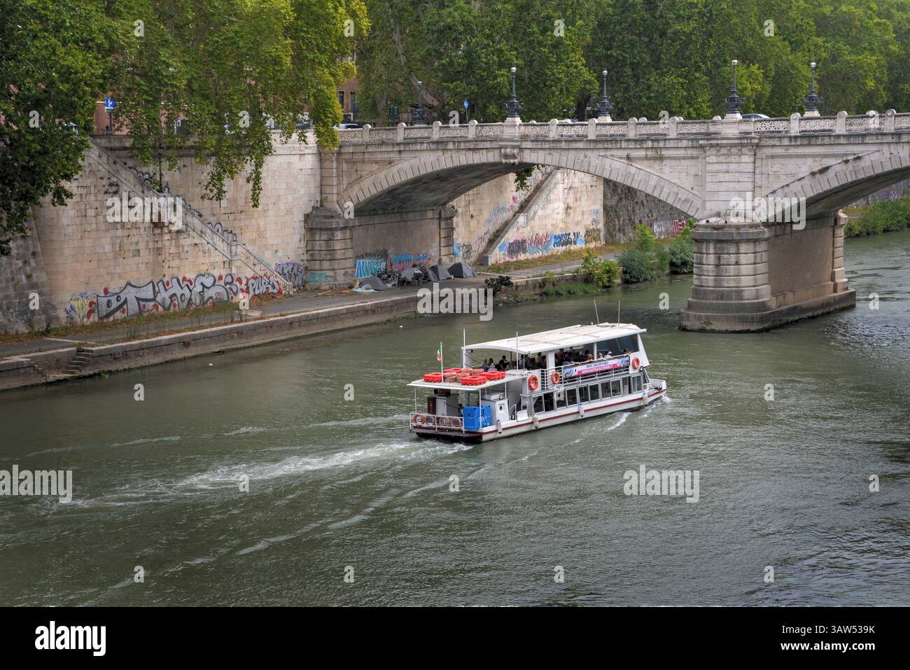 Rom, Italien - 8. September 2024: Touristenboot auf dem Tiber in Rom, vorbei an einer historischen Brücke, umgeben von Graffiti-bedeckten emba Stockfoto