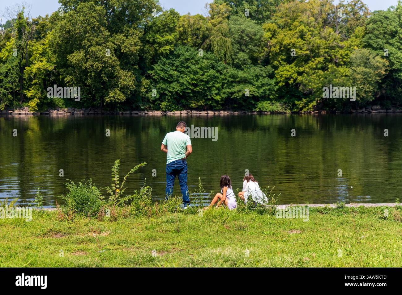 Ein Mann und zwei Kinder fischen im Schuylkill River. Der Mann steht und die Kinder sitzen auf dem Gras. Die Szene ist friedlich und entspannt Stockfoto