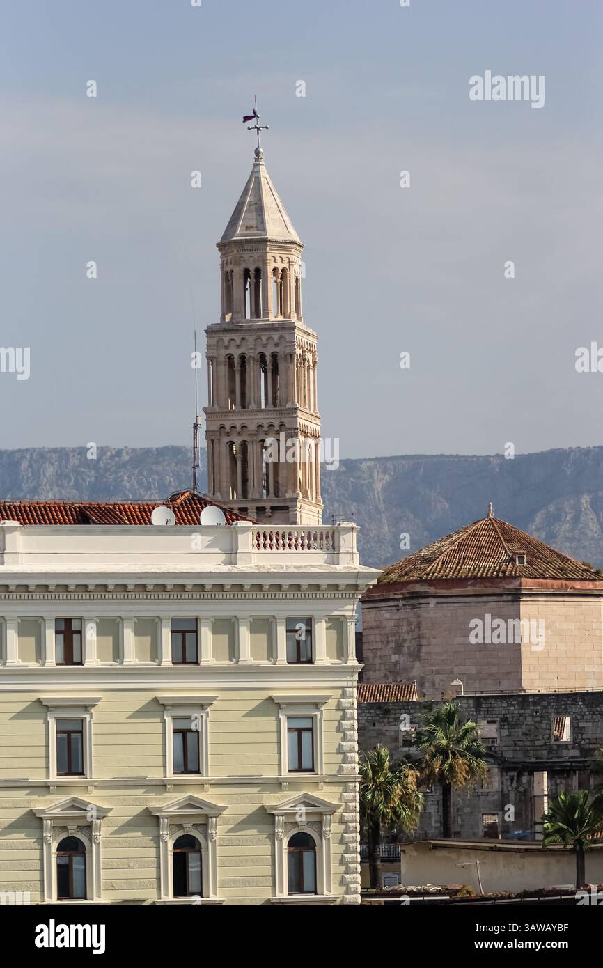 Blick auf einen aufwendig gestalteten Glockenturm, der sich vor einem entfernten Bergmassiv unter einem ruhigen blauen Himmel erhebt, umgeben von bezauberndem Histo Stockfoto