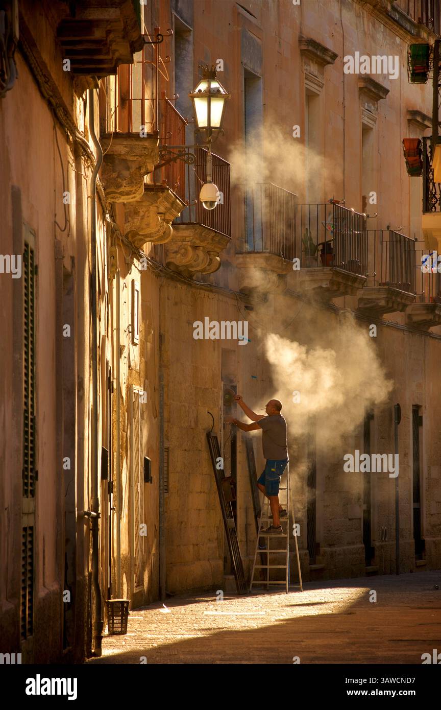 Italienischer Arbeiter auf einer Leiter, der an einer Tür arbeitet, sein Winkelschleifer weht eine Wolke aus Staub, hinterleuchtet von der goldenen Morgensonne. Lecce, Apulien, Süditalien Stockfoto