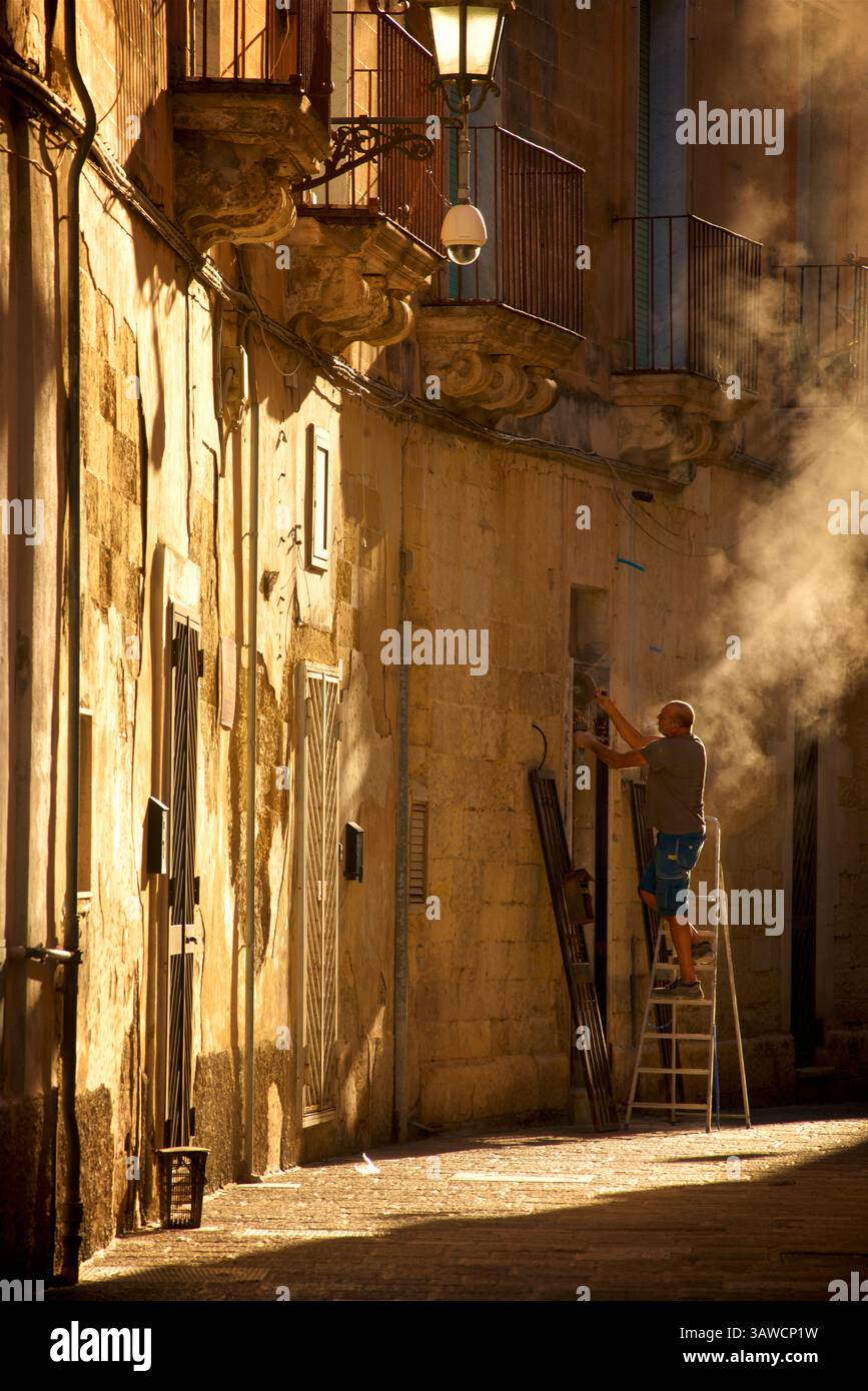 Italienischer Arbeiter auf einer Leiter, der an einer Tür arbeitet, sein Winkelschleifer weht eine Wolke aus Staub, hinterleuchtet von der goldenen Morgensonne. Lecce, Apulien, Süditalien Stockfoto