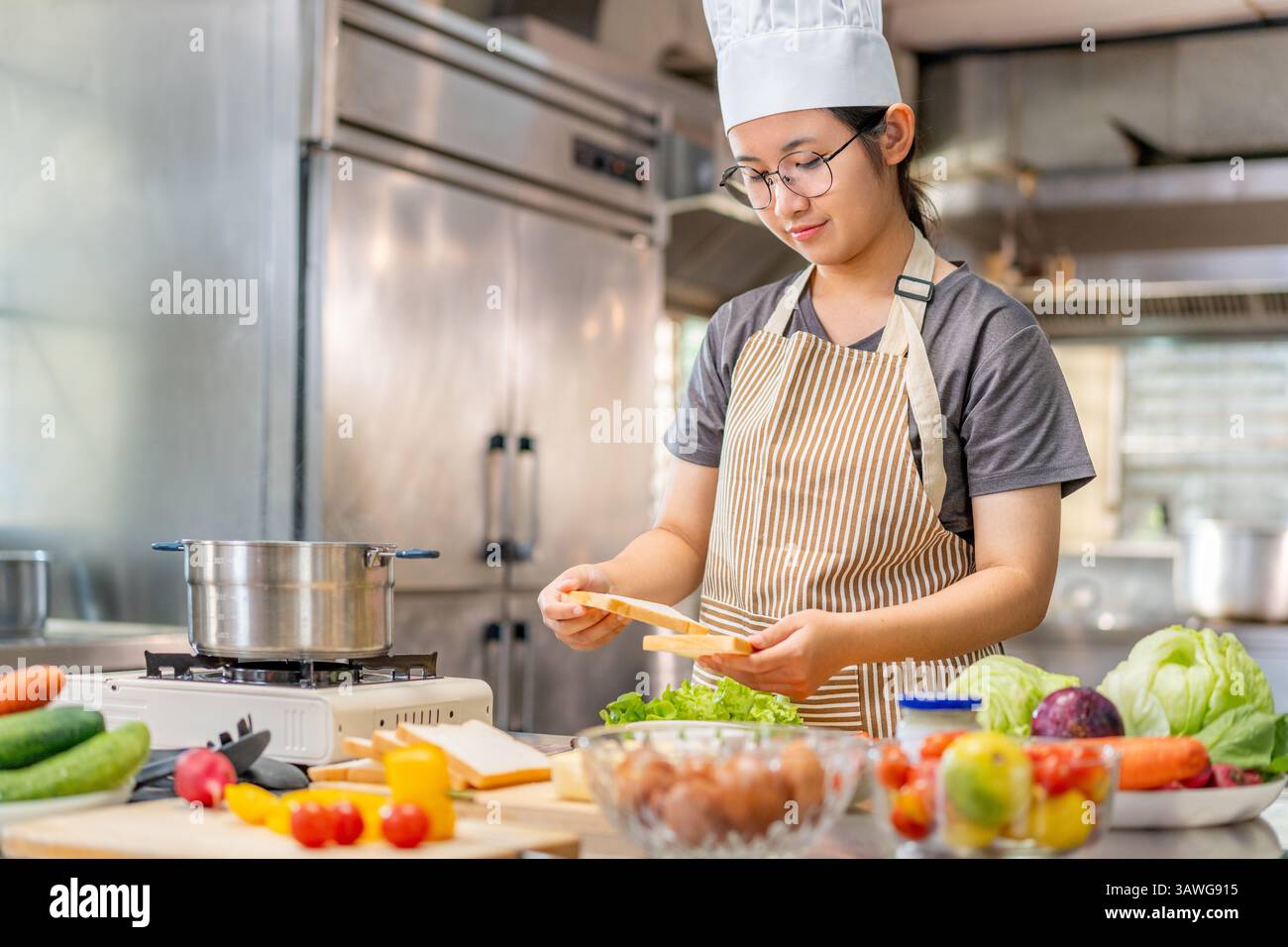 Eine fröhliche junge Küchenchefin, die eine gestreifte Schürze und einen weißen Kochhut trägt, sieht man, wie sie ein frisches Sandwich in einer kommerziellen Küche zubereitet. Umgeben von fres Stockfoto