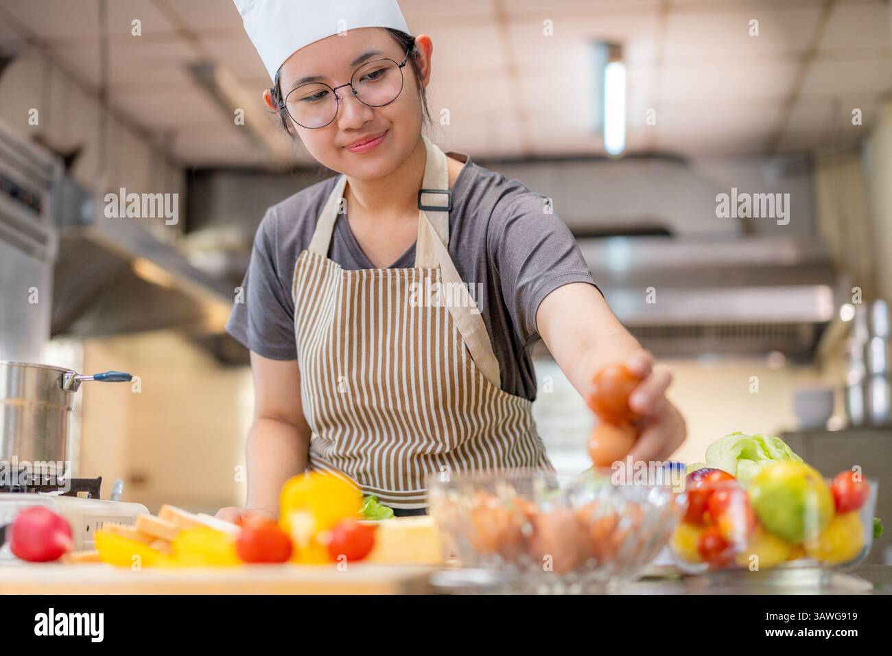 Junge Küchenchefin mit Schürze und Hut bereitet frische Zutaten in einer kommerziellen Küche zu. Sie lächelt, während sie ein Ei auswählt, umgeben von bunten Stockfoto