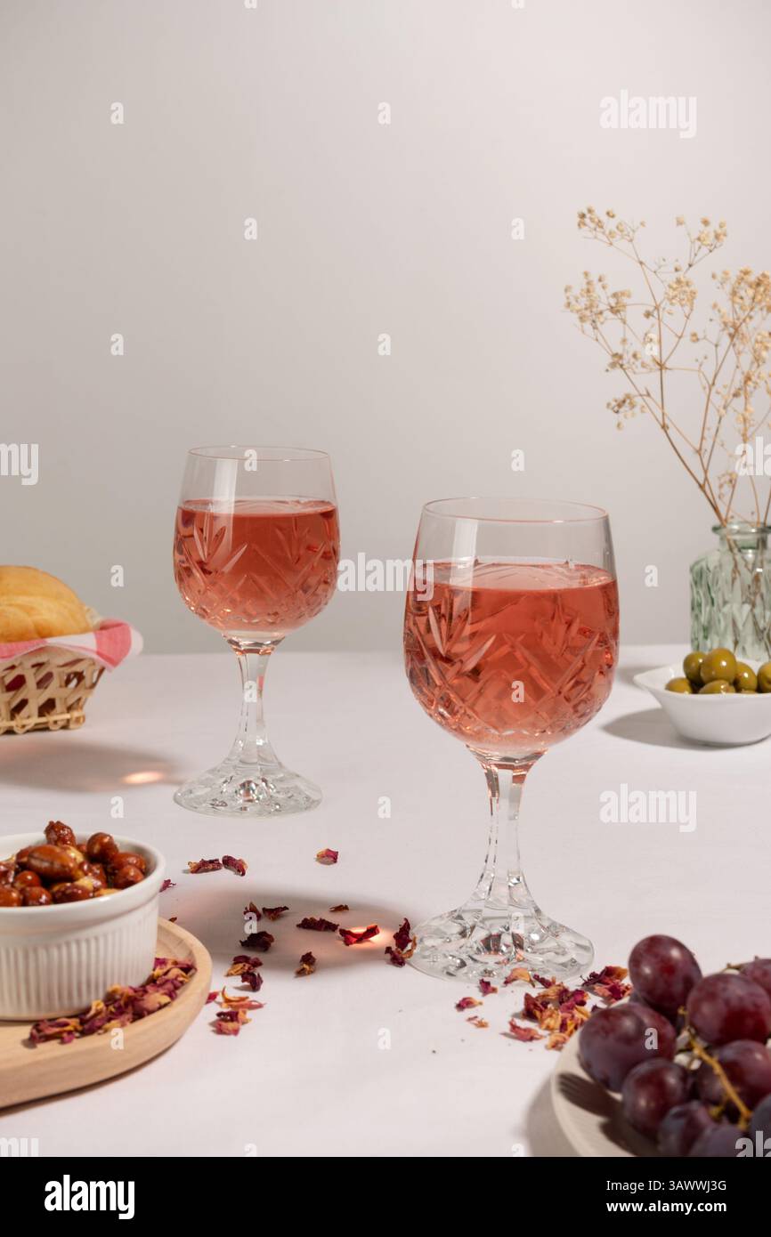 Zwei Gläser Rosenwein auf einer weißen Tischdecke. Auf dem Tisch befindet sich eine kleine Schüssel mit Erdnüssen, roten Trauben, einem Brotkorb und einer grünen Vase. Stockfoto