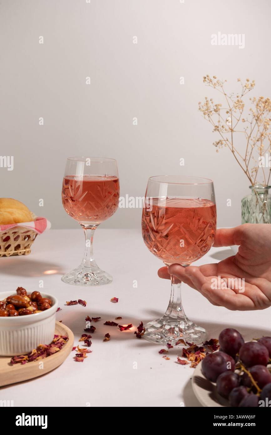 Zwei Gläser Rosenwein auf einer weißen Tischdecke, eine Hand nimmt eines davon. Auf dem Tisch steht eine kleine Schüssel mit Erdnüssen, roten Trauben. Stockfoto