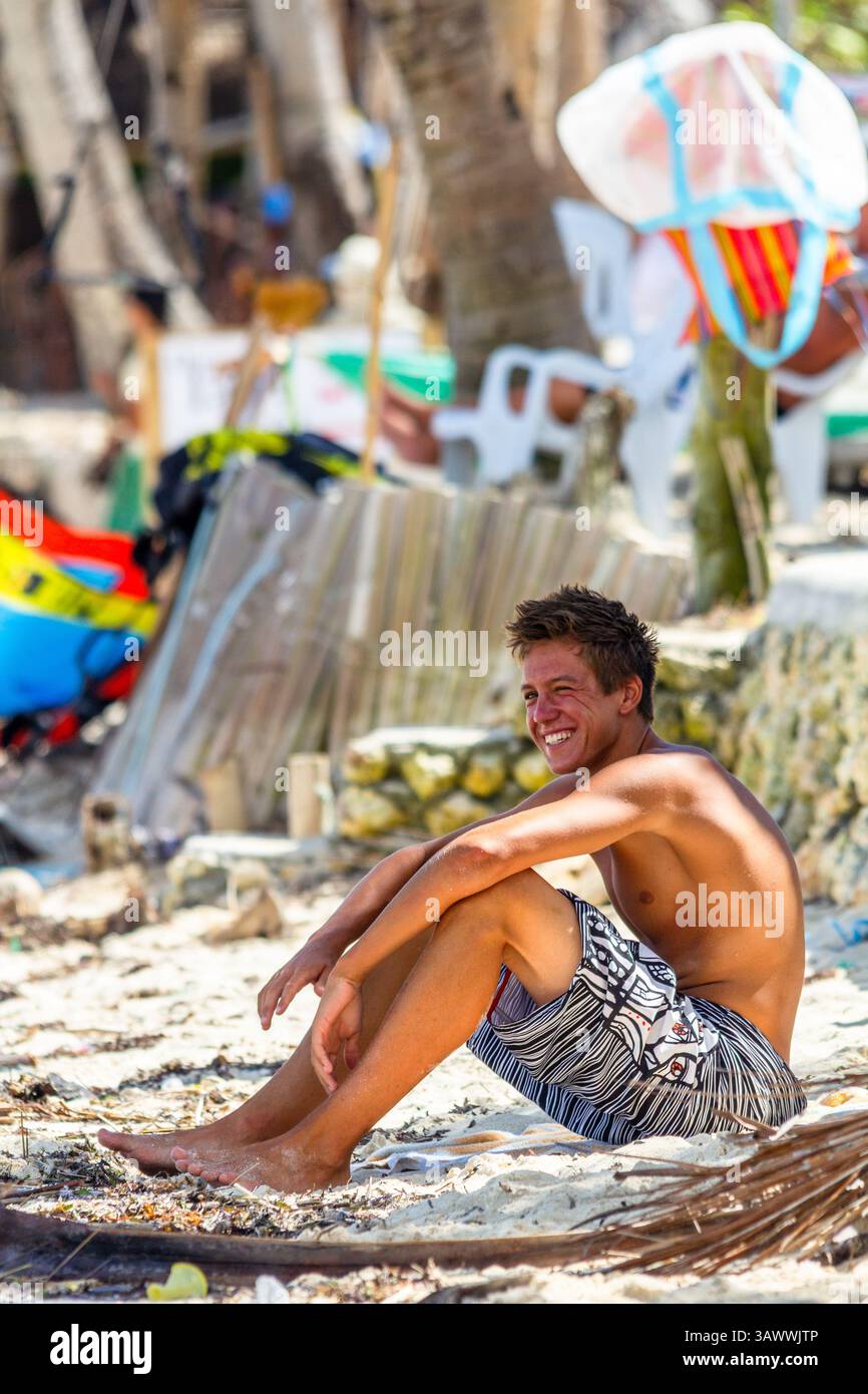 Ein lächelnder junger weißer Mann sitzt auf dem pulverförmigen weißen Sand von Boracay Island, Philippinen und genießt die tropische Strandatmosphäre Stockfoto