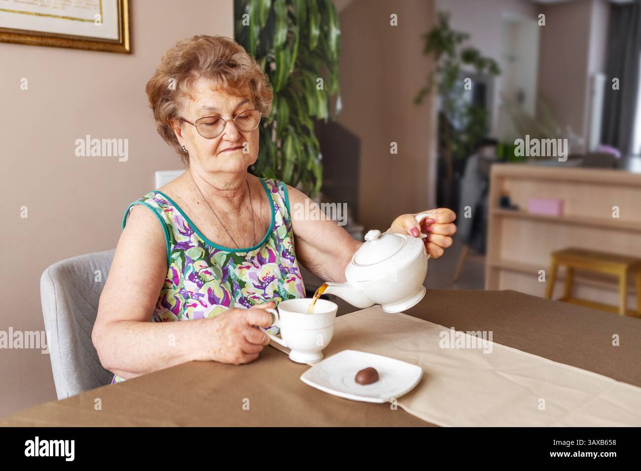 Seniorin mit kurzen Haaren und Gläsern, die allein zu Hause sitzen, Tee mit süßen Süßigkeiten auf dem Tisch trinken, ungesunde Essgewohnheiten und Risiken des Zuckerkonsums Stockfoto