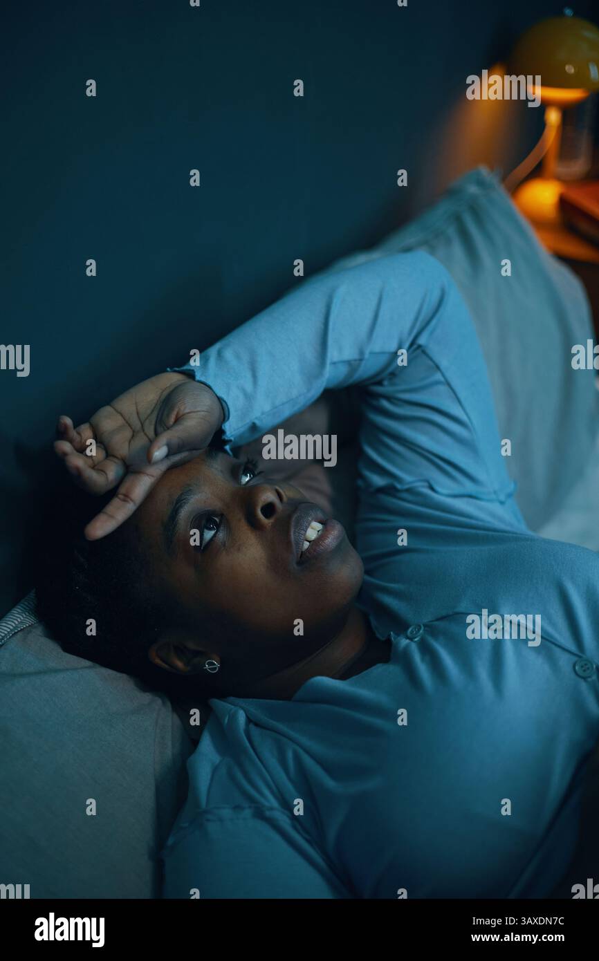 Person, die unter schwachem Licht auf dem Bett liegt und Ausdruck tiefe Gedanken anzeigt. Die orangefarbene Lampe leuchtet im Hintergrund sanft und trägt zur Kontemperierung bei Stockfoto