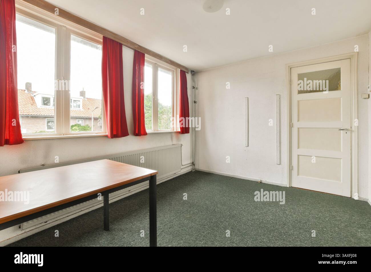 Ein helles, unmöbliertes Zimmer mit großen Fenstern mit roten Vorhängen. Der Raum hat ein minimalistisches Design und neutrale Wände, ideal für verschiedene Zwecke. Stockfoto