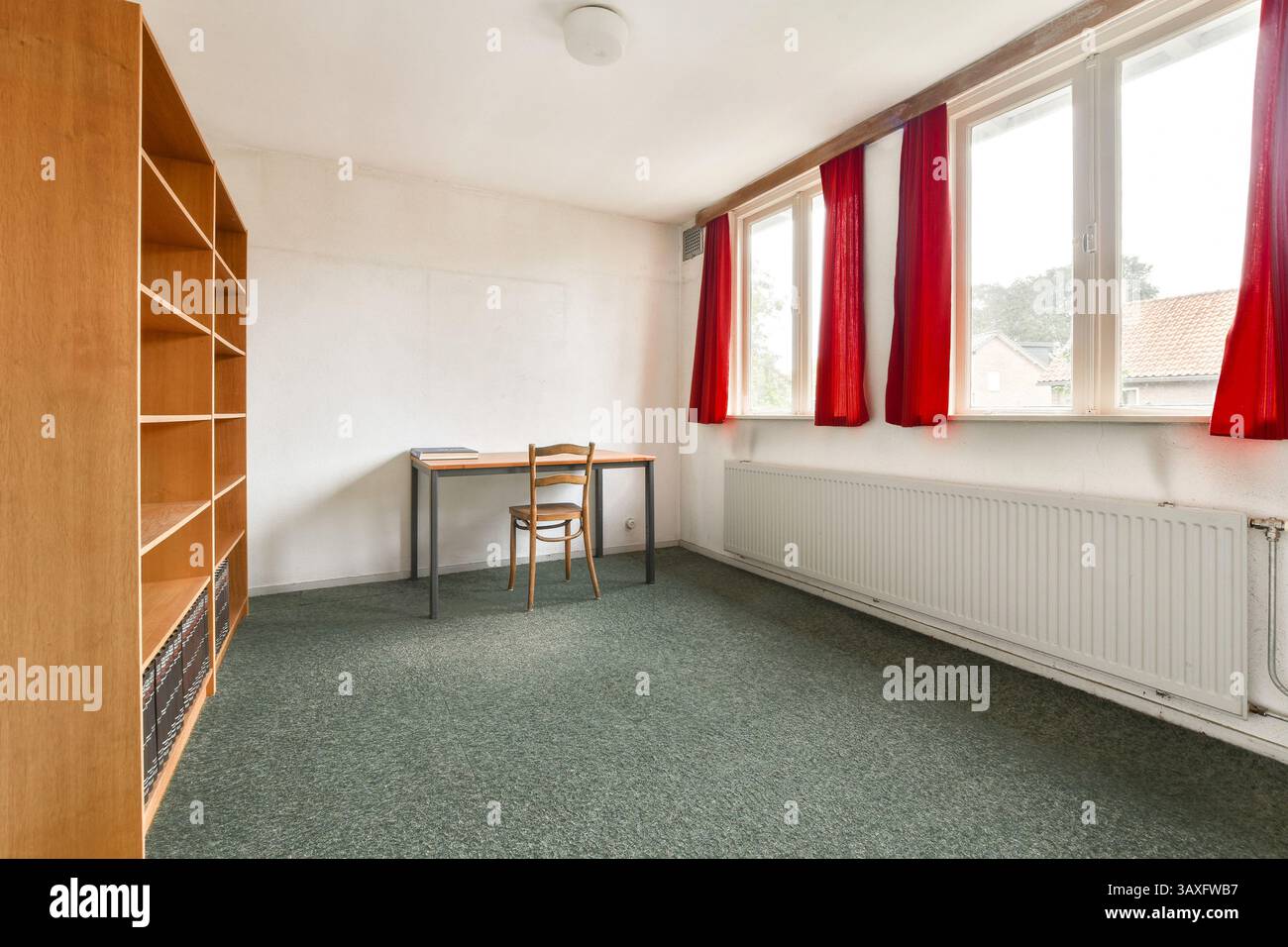 Ein einfacher und sauberer Innenraum mit einem Holztisch, einem Stuhl und hellroten Vorhängen im Kontrast zu neutralen Wänden und Teppichen. Stockfoto