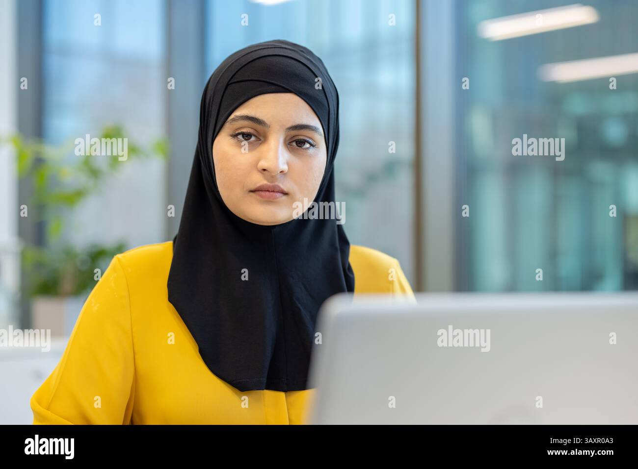 Eine Frau mit schwarzem Hijab und gelber Bluse posiert mit ernstem Ausdruck in einem Büro. Der Laptop deutet an, dass sie arbeitet. Stockfoto