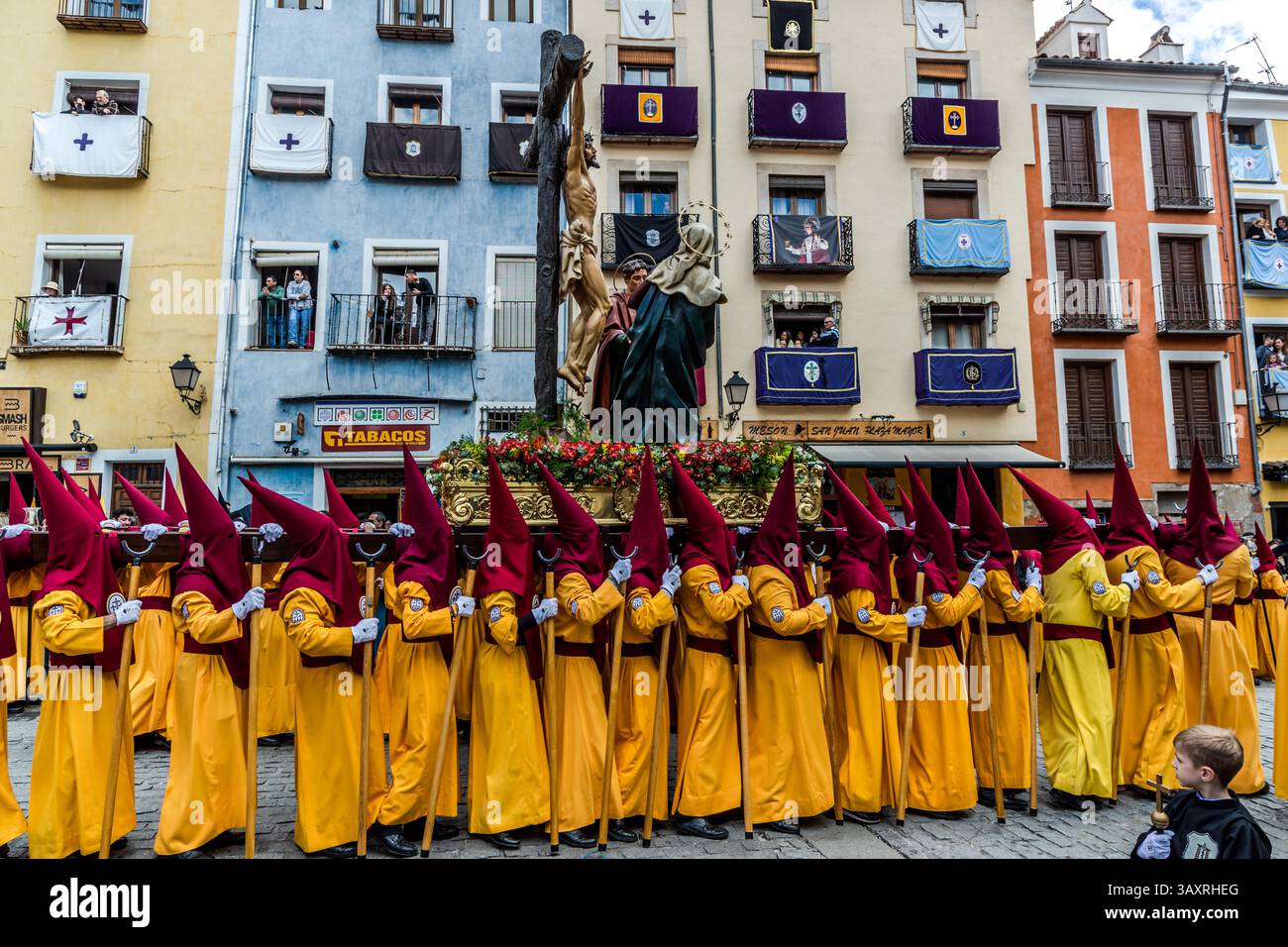 Träger (Costaleros) der ehrwürdigen Hermandad del Santísimo Cristo de la Agonía tragen den Paso Santísimo Cristo de la Agonía (Federico Coullaut-Valera) auf ihren Schultern durch Cuenca, Castilla-La Mancha, Spanien. Lila und blaue Bruderschaftsbanner und ein gerahmtes Porträt hängen von farbenfrohen Fassaden, während Zuschauer von Fenstern und Balkonen aus zuschauen. Während der Semana Santa in Cuenca nehmen 33 Hermandades an elf Prozessionen durch die Stadt während der Heiligen Woche teil. Stockfoto