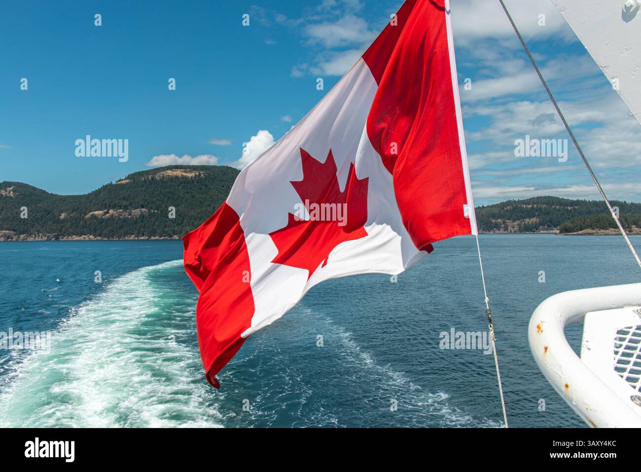 Rot-weiße kanadische Flagge, die auf einer fahrenden Fähre im Wind weht, mit bewaldeten Küsteninseln im Hintergrund. Stockfoto