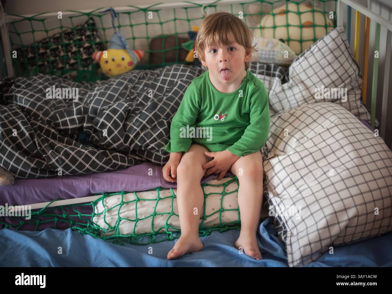 Ein kleines Kind in einem grünen Hemd sitzt auf einem gemütlichen Bett mit bunten Kissen und Spielzeug, wobei es spielerisch seine Zunge herausstreckt und sich in einem ch entspannt Stockfoto