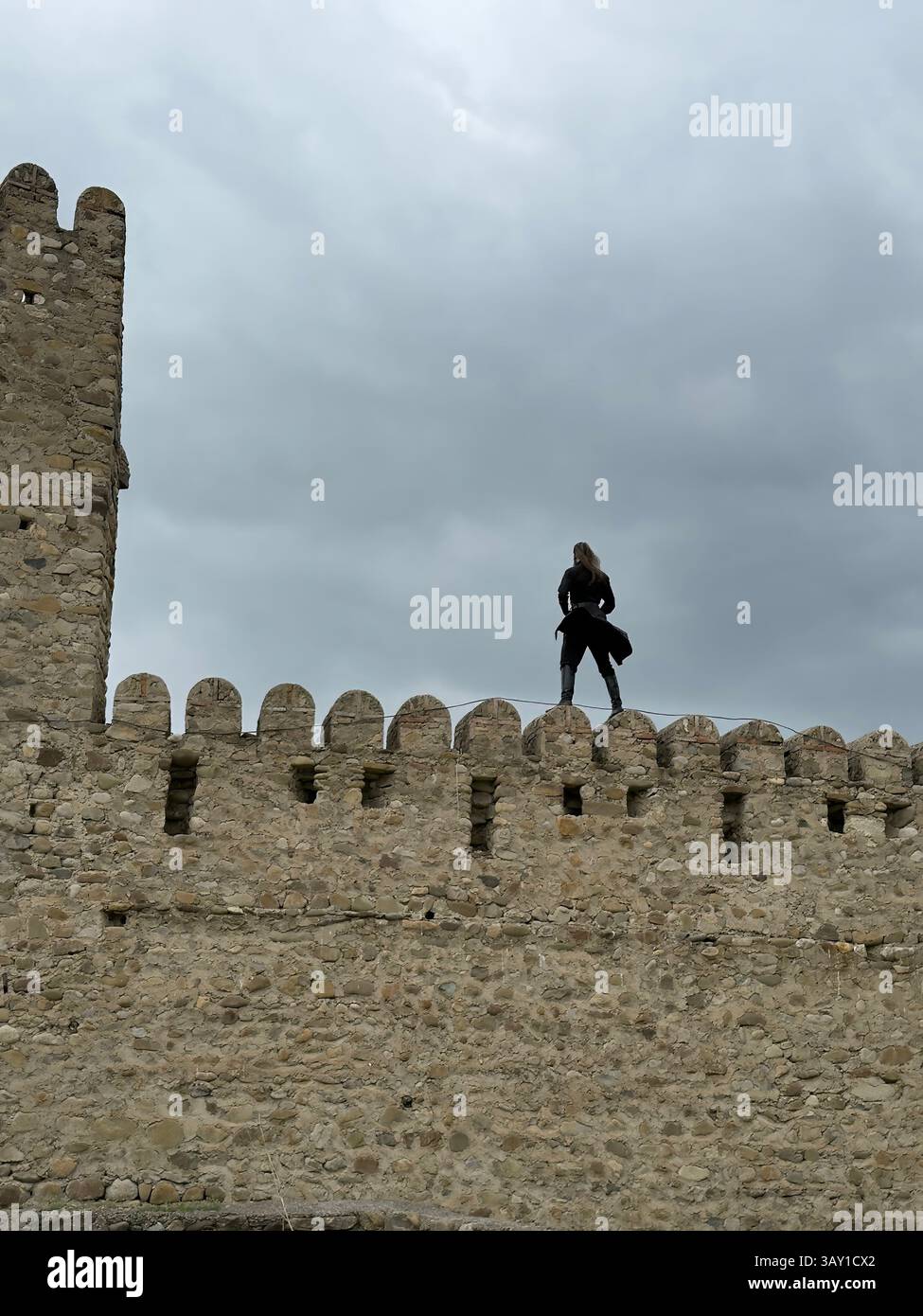 Mann, der auf einer alten Steinfestungsmauer mit Zinnen vor bewölktem Himmel steht, historische Architektur und mittelalterliche Befestigungen. Reisen, Geschichte, - Smartphone-aufgenommenes Stockfoto