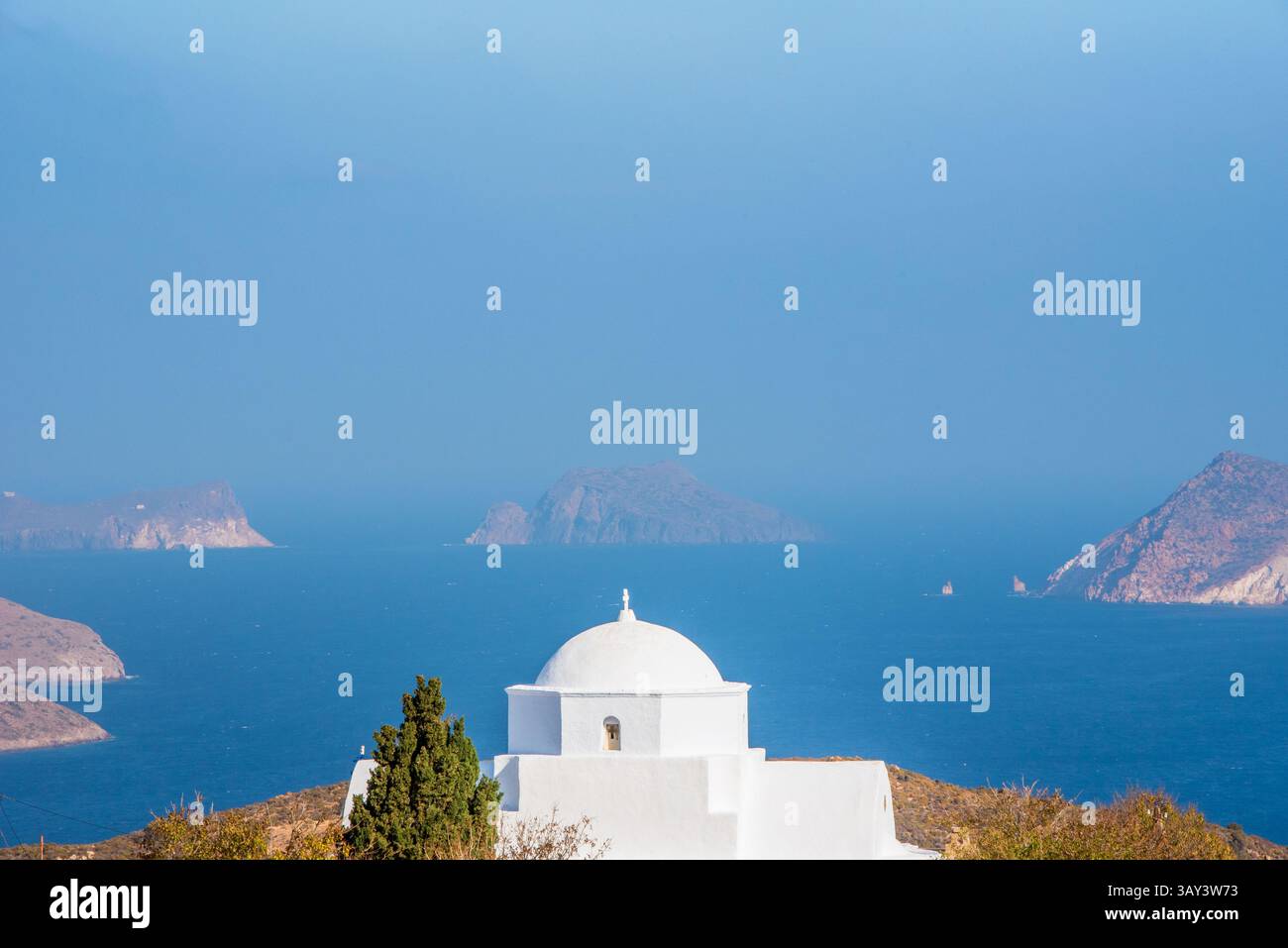 Eine wunderschöne weiße Kirche mit Kuppel überblickt das ruhige blaue Meer und die fernen Inseln auf Milos, Griechenland. Die ruhige Aussicht verkörpert den Charme des GRE Stockfoto