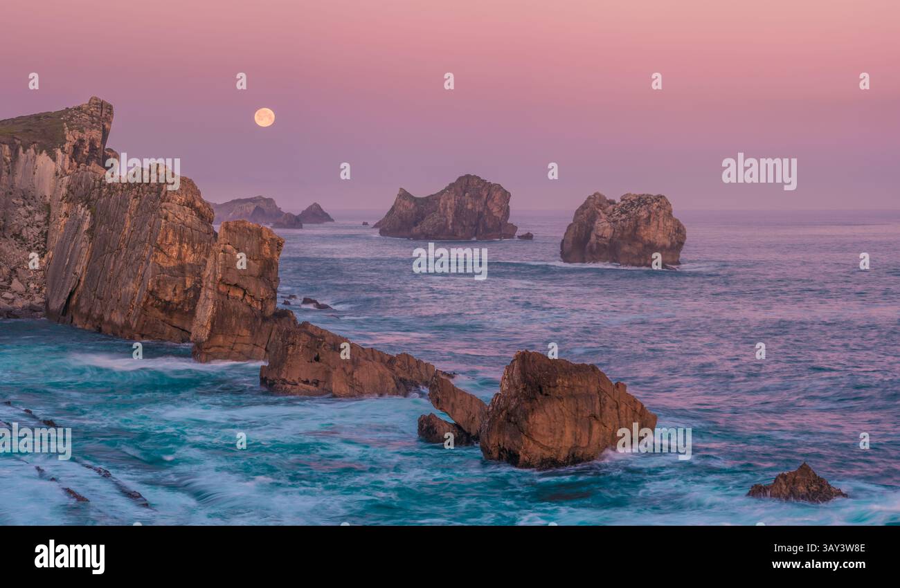 Ein ruhiger Blick auf zerklüftete Seestapel und Klippen im sanften Morgenlicht am Strand von Arnia, Costa Quebrada, mit Vollmond über der ruhigen A Stockfoto