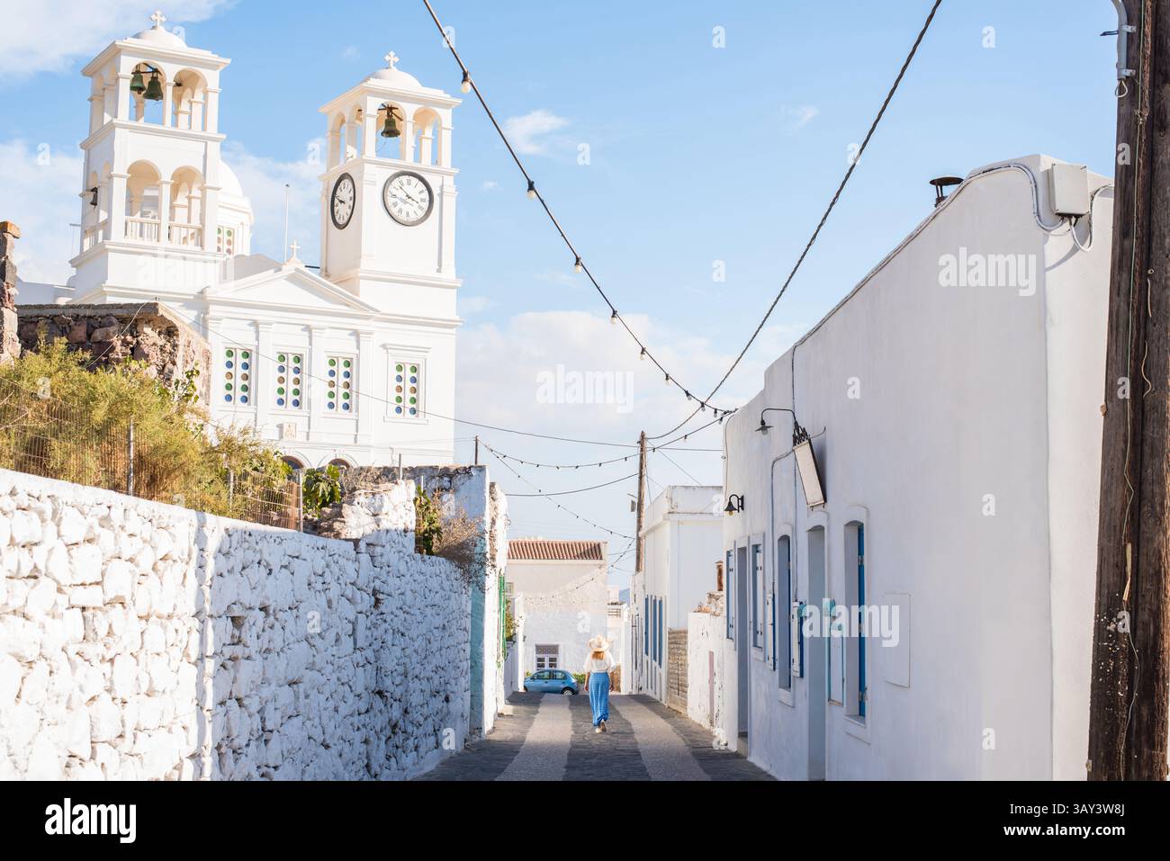 Frau, die eine malerische Straße in Trypiti, Milos Island, erkundet, mit klassischer griechischer Architektur mit einer weißen Kirche, Glockentürmen und einem Kopfsteinpflaster Stockfoto