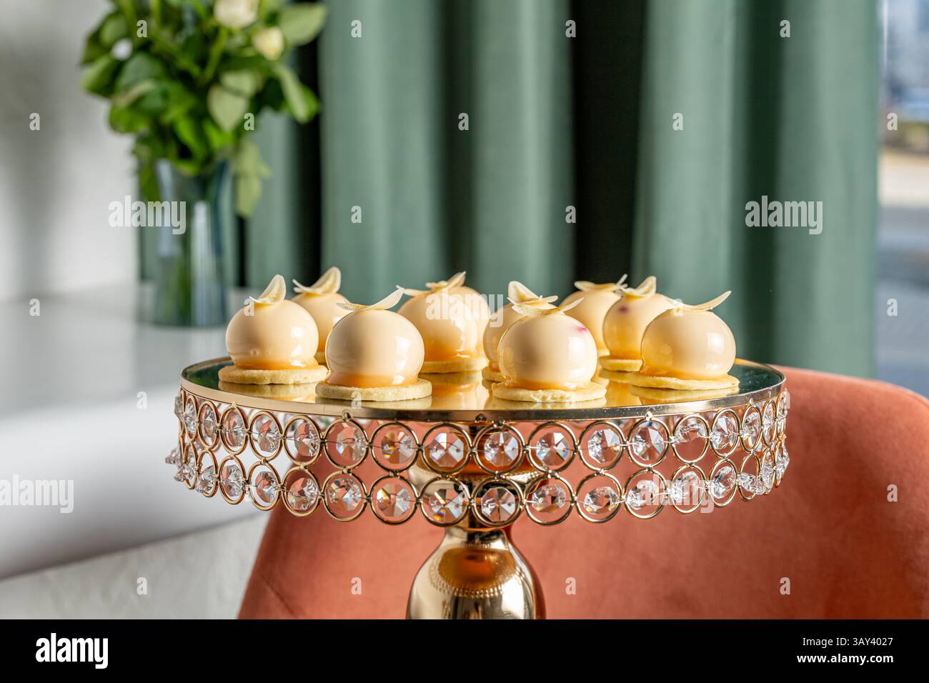 Eine Auswahl an exquisiten Vanillemousse-Desserts befindet sich auf einem kunstvollen Kristallständer vor einem Hintergrund mit eleganten Vorhängen und Blumendekorationen Stockfoto