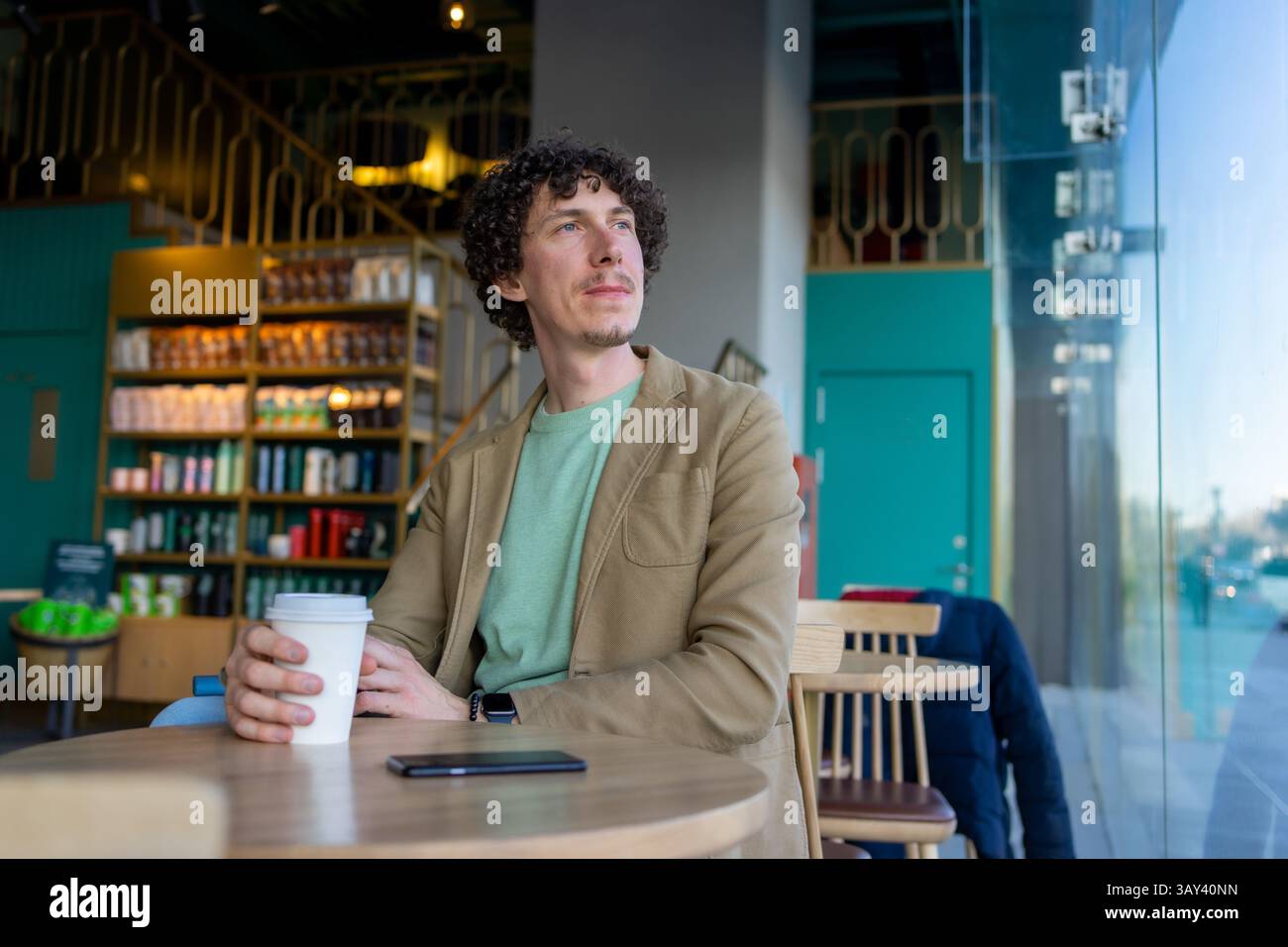 Ein aufmerksamer Mann sitzt in einem modernen Café, hält eine Kaffeetasse und blickt aus dem Fenster. Helles Interieur, Regale voller Produkte und eine entspannte Atmosphäre Stockfoto