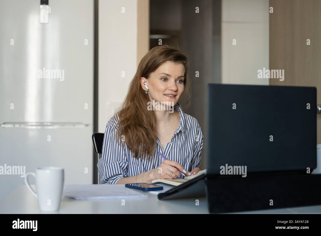 Eine Frau sitzt an einem Tisch und arbeitet von zu Hause aus. Sie schreibt in einem Notizbuch, während sie sich auf ihren Laptop-Bildschirm konzentriert, mit einem Becher und einem Telefon in der Nähe. Stockfoto