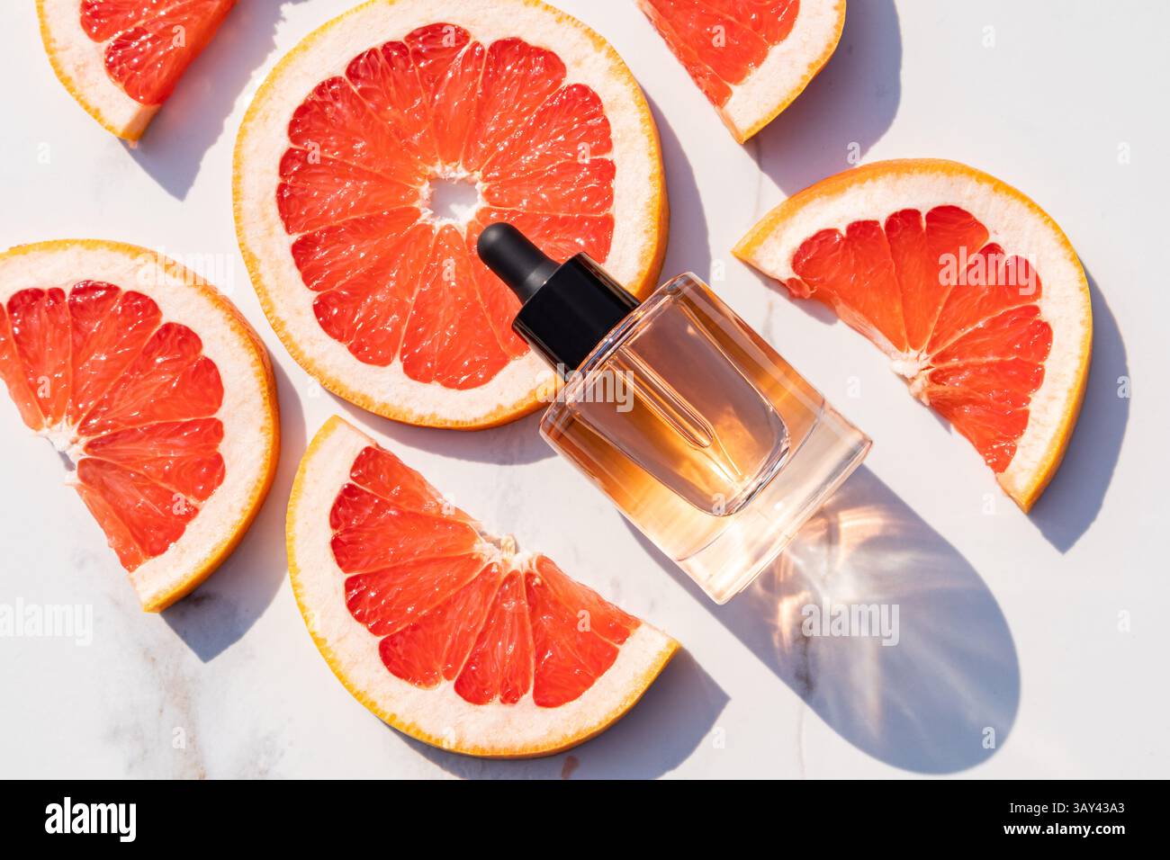 Helle Grapefruitscheiben umgeben eine elegante Tropfflasche mit ätherischem Öl zur Hautpflege auf einer Marmoroberfläche. Sonnenlicht erhellt einen frischen, lebendigen Kamm Stockfoto