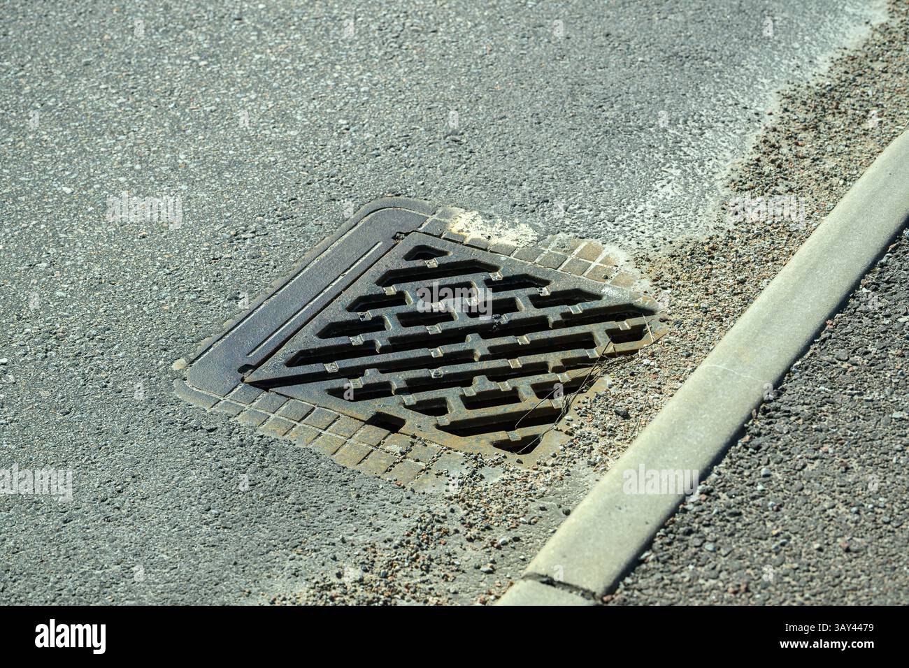 Abflussabdeckung auf einer Betonstraße, die sich mit dem umgebenden Schotter verschmilzt. Die Bordsteinkante ist klar definiert und hebt die städtische Infrastruktur hervor Stockfoto