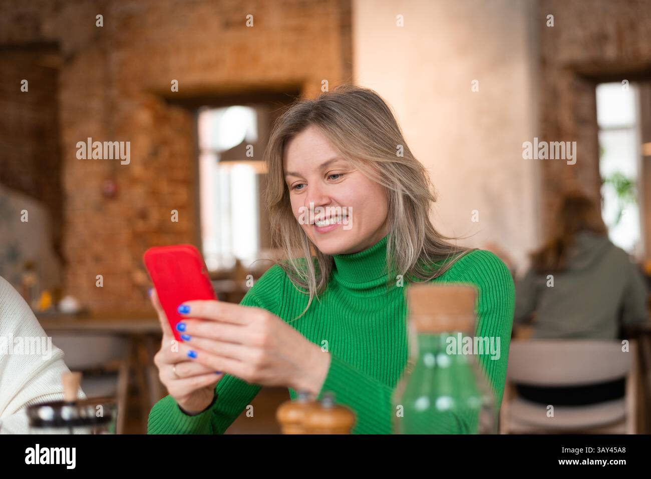 Eine Frau in einem grünen Pullover lächelt, während sie ihr Smartphone in einem gemütlichen Café ansieht. Sie genießt ein friedliches Frühstück, umgeben von einem warmen Atmo Stockfoto