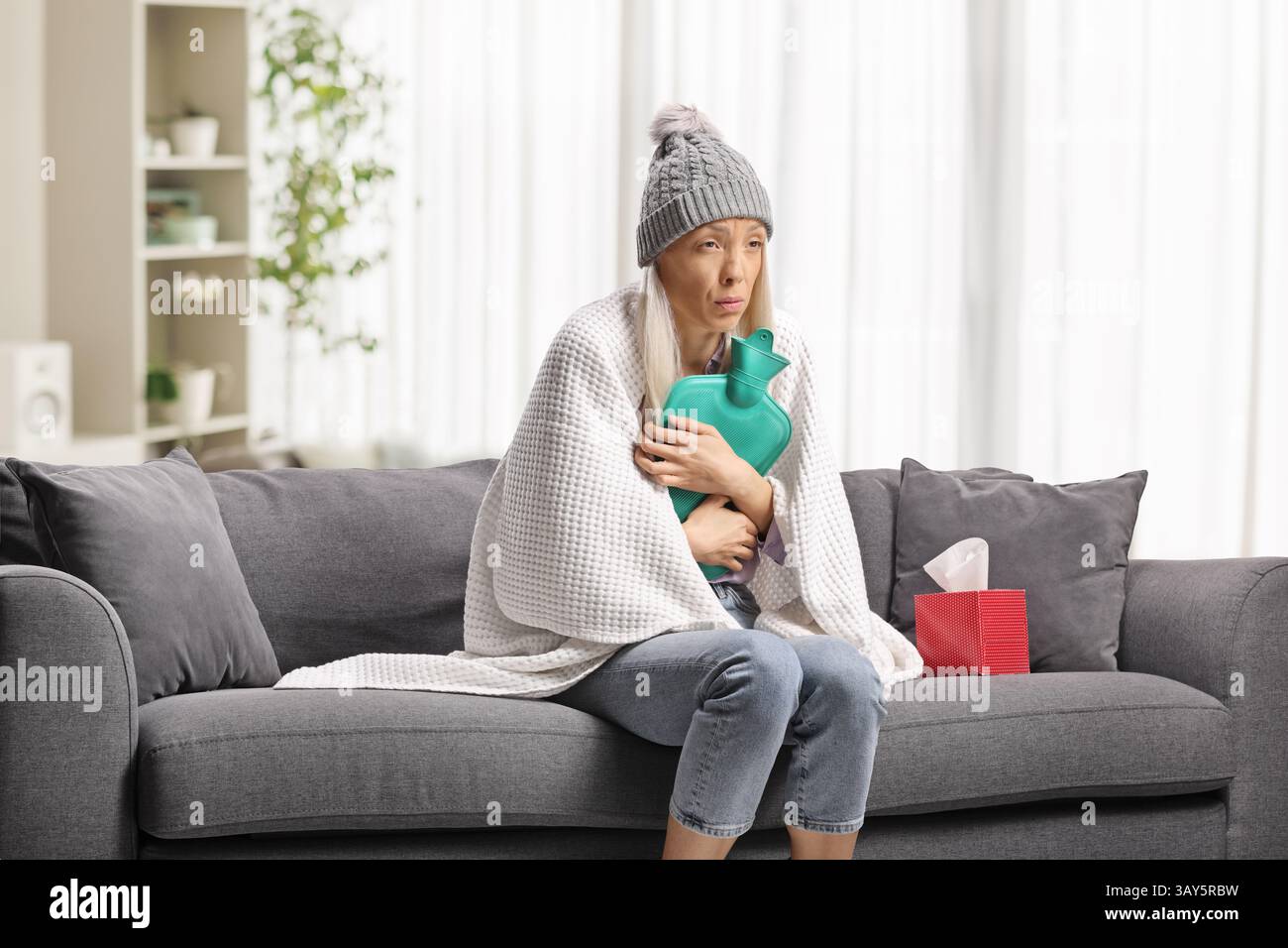 Frau, in Decke gewickelt, auf einem Sofa sitzend und mit einem Körperwärmer in der Hand Stockfoto