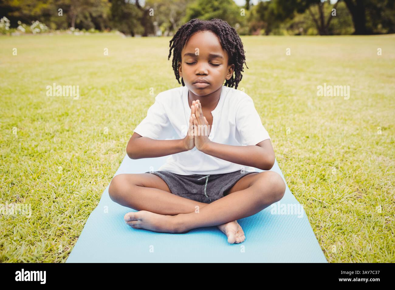 Afroamerikanischer Junge, der auf blauer Yogamatte auf einem grasbewachsenen Feld meditiert, Ruhe und Konzentration genießt Stockfoto