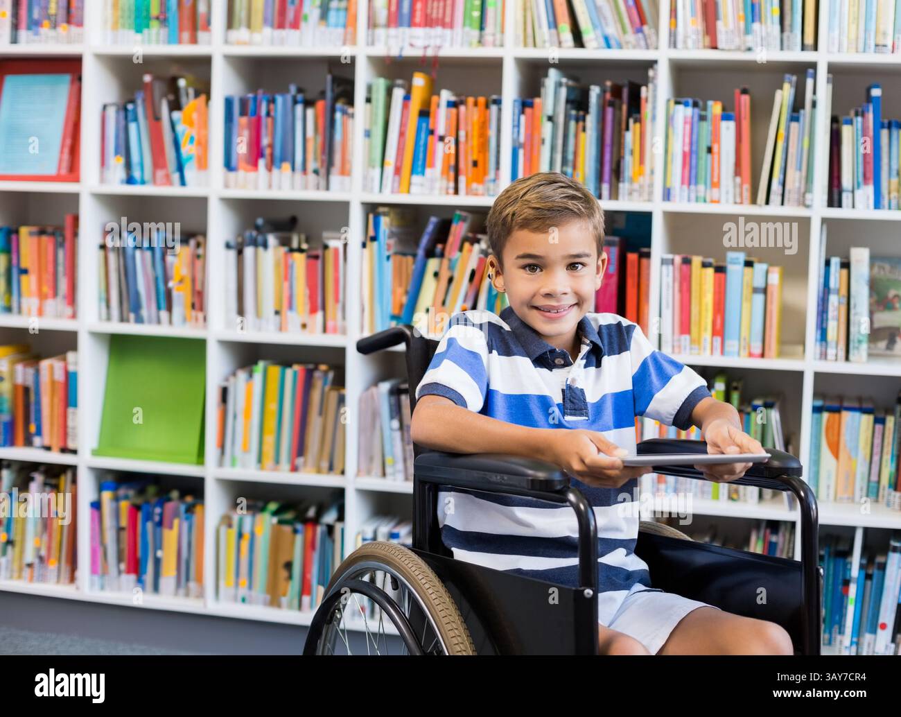 Schuljunge sitzt im Rollstuhl in der Bibliothek und hält ein Tablett mit bunten Bücherregalen dahinter Stockfoto