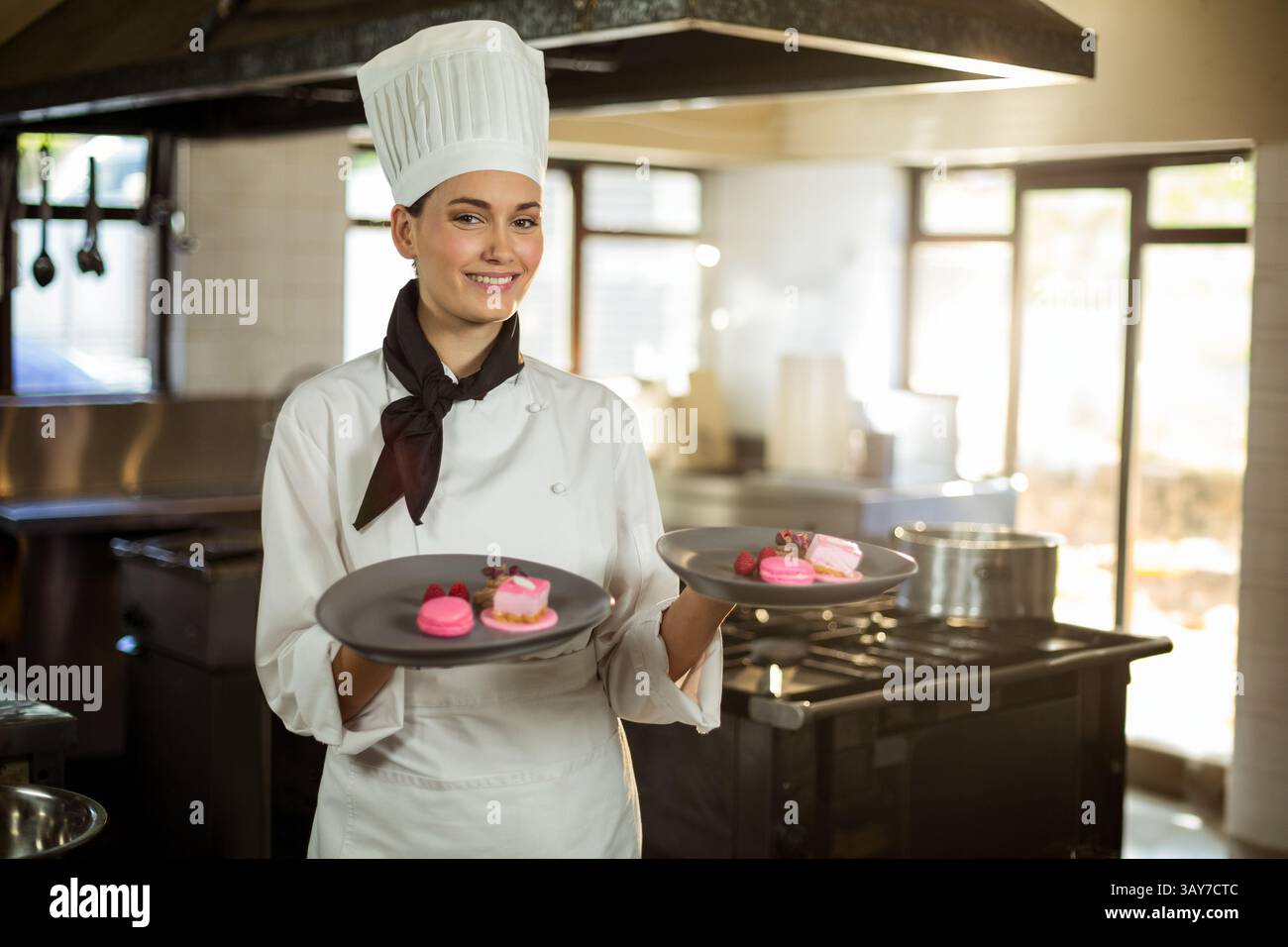 Junge Küchenchefin hält zwei Teller mit rosa Macarons in der kommerziellen Küche und präsentiert Gebäck Stockfoto