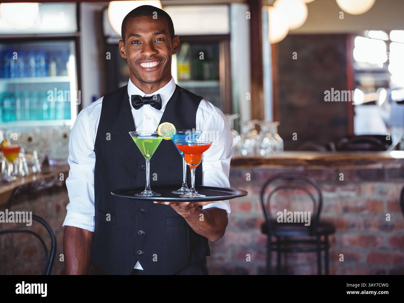 Afroamerikanischer Mann serviert Cocktails an der Bar und hält ein rundes Tablett mit grünen, blauen und roten Getränken Stockfoto