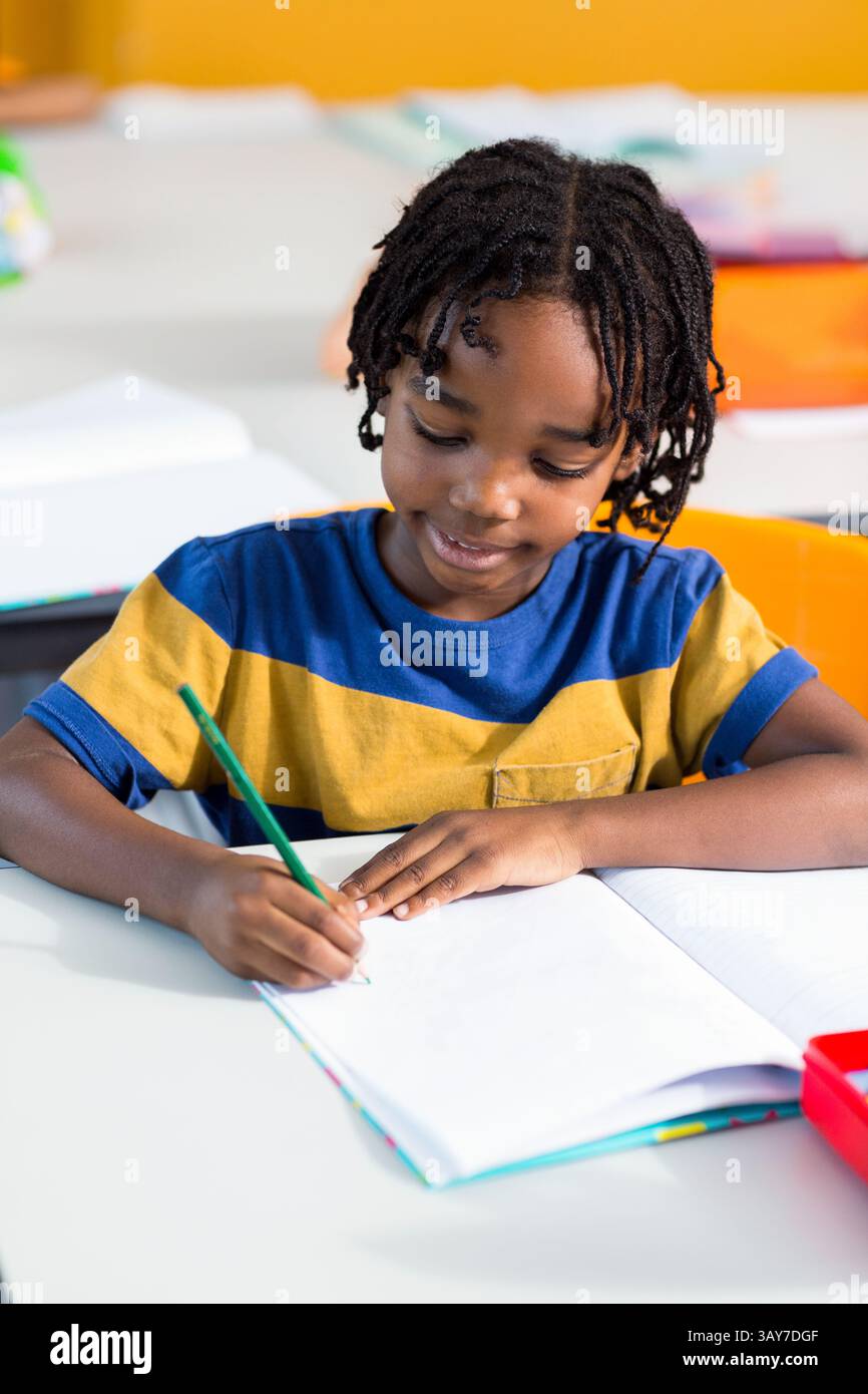 Afroamerikanischer Junge, der in offenem Notizbuch am Schreibtisch im Klassenzimmer schreibt, mit grünem Bleistift und rotem Tablett Stockfoto